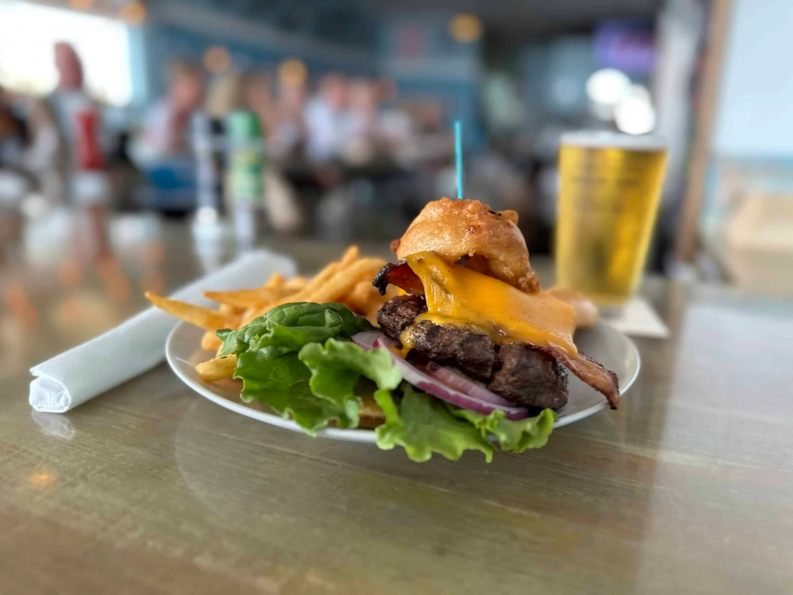 Bacon cheeseburger topped with an onion ring, with fries and a draft beer at the bar